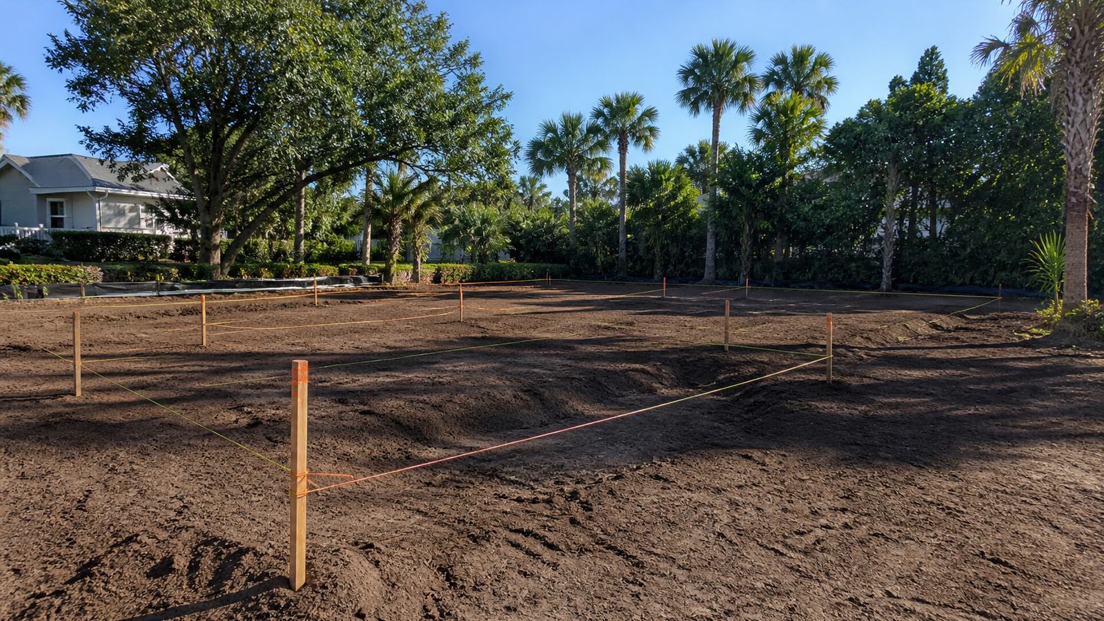 Coastal residential lot laid out with survey stakes, string lines, graded footprint, and mature vegetation