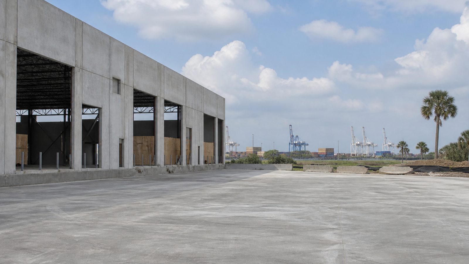 Wide concrete warehouse shell beside an empty loading apron with distant port cranes