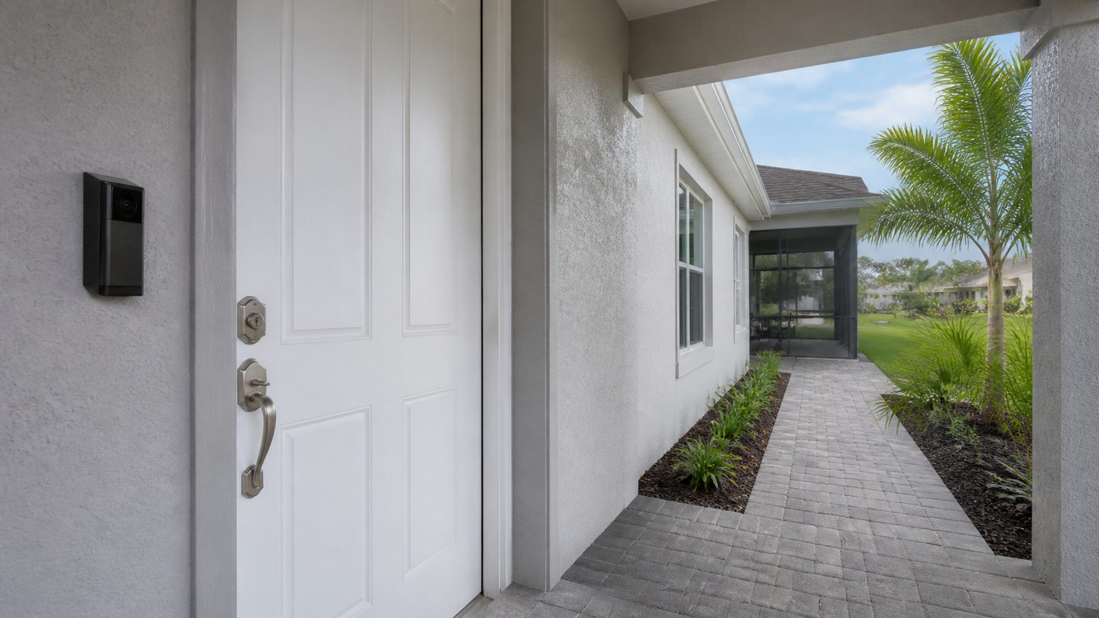 Black video doorbell beside a white stucco entry with a paver walk and palms